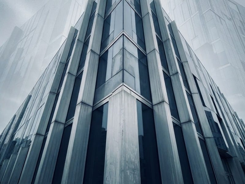 A low-angle view of the corner of a modern building featuring vertical concrete pillars and glass panels against a foggy sky.